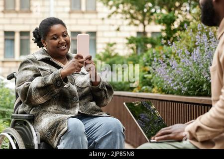 Portrait of smiling black woman in wheelchair taking mobile photo in city outdoors, copy space Stock Photo