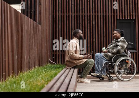 Graphic side view shot of young woman in wheelchair talking to friend or colleague outdoors in city park, copy space Stock Photo
