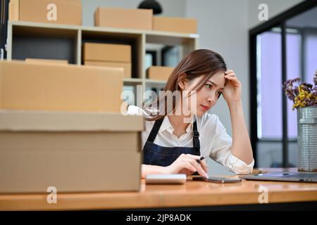 Attractive young Asian female entrepreneur or e-commerce business startup in the office perplexed by her online store website. Stock Photo