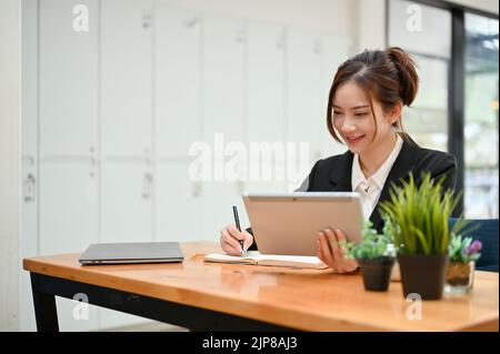 Attractive young Asian female assistant or secretary working at her office desk, using tablet while writing something on her notebook. Stock Photo