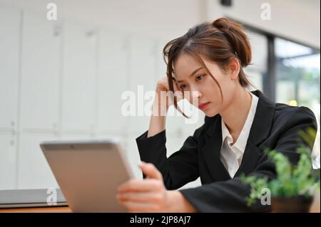 Thoughtful young Asian businesswoman using tablet, contemplating a business strategy and concentrating on her project. Stock Photo