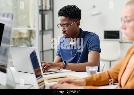 African young programmer concentrating on his online work, he typing on laptop while sitting at table with colleagues at office Stock Photo