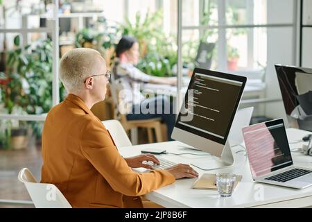 Rear view of female developer with short hair sitting at her workplace with computer monitor and typing codes Stock Photo
