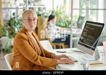 POrtrait of mature female programmer looking at camera while sitting at table with computer and working with codes Stock Photo