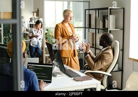 African programmer telling his idea to woman while she making notes in her notepad during meeting at office Stock Photo