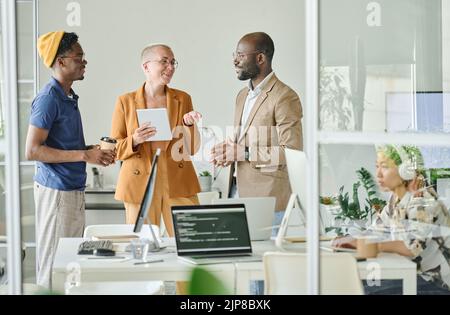 Group of programmers discussing online project in team while standing at office Stock Photo