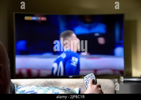 A  hand presing a button on the remote control in front of a television Stock Photo
