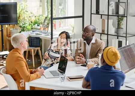 Group of programmers sitting at table and discussing together new app of VR glasses during meeting at office Stock Photo
