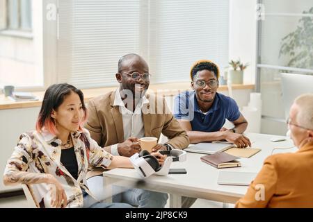 Group of multiethnic developers sitting at table and working with app of virtual reality glasses during teamwork at office Stock Photo