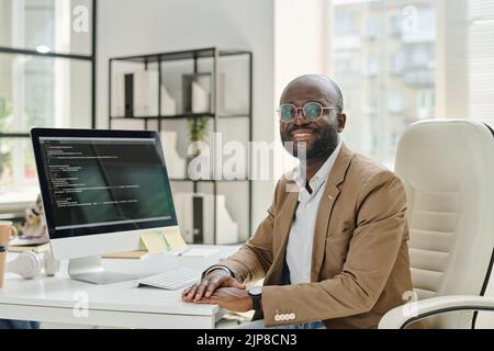 Portrait of African developer in eyeglasses smiling at camera while sitting at his workplace with computer monitor with codes on screen Stock Photo