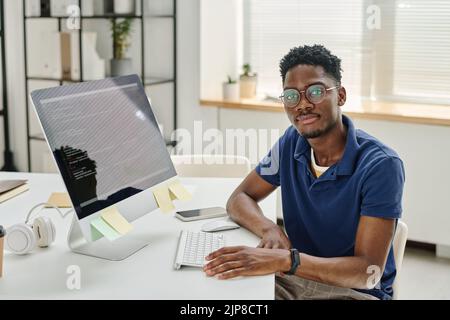 Portrait of African young developer in eyeglasses looking at camera while sitting at his workplace with computer monitor with codes on screen Stock Photo