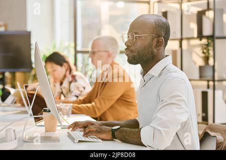 African programmer in eyeglasses typing on keyboard writing codes on computer while sitting at table with his colleagues in background Stock Photo