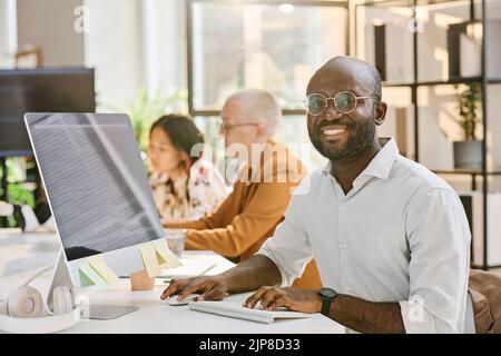 Portrait of African programmer in eyeglasses smiling at camera while working on computer at table at office Stock Photo