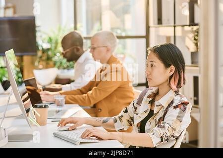 Asian young programmer sitting at table and using computer to type codes with her colleagues in background Stock Photo