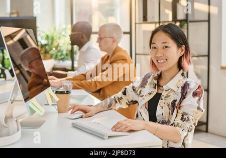 Portrait of Asian female programmer smiling at camera while sitting at table in front of computer with her colleagues Stock Photo