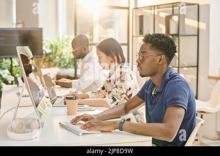 African young programmer in eyeglasses concentrating on his work on computer sitting at table with his colleagues at office Stock Photo