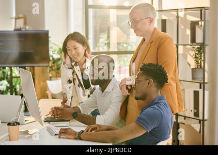 Multiethnic group of programmers looking at computer monitor and discussing software in teamwork at office Stock Photo