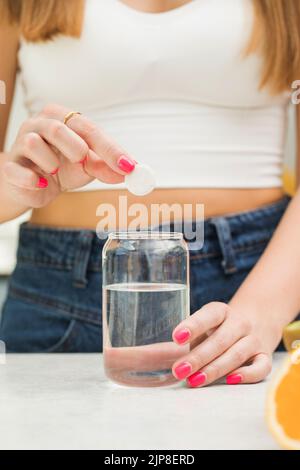 A woman throws a white round pill into a glass of water. Pain reliever, complex vitamins for women's health. Stock Photo