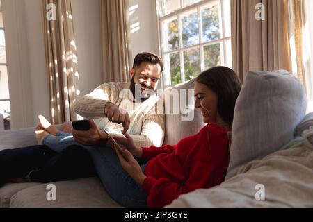 Happy caucasian couple sitting on couch in living room laughing, using smartphone and tablet Stock Photo