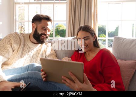Happy caucasian couple sitting on couch in living room laughing, looking at tablet Stock Photo