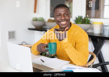 Smiling african american man using laptop at home, talking on smartphone and drinking coffee Stock Photo