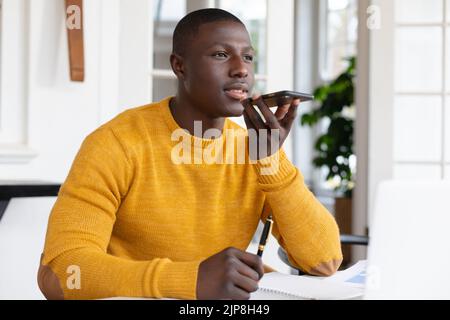 African american man at home writing notes and talking on smartphone Stock Photo