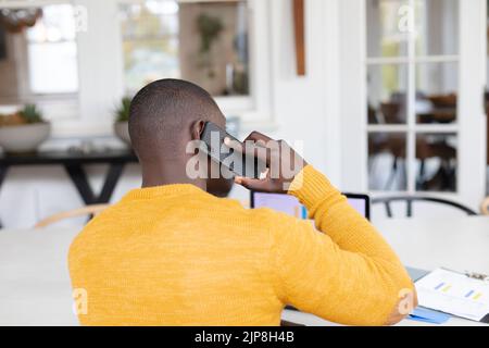 Rear view of african american man using laptop at home and talking on smartphone Stock Photo