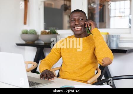 Smiling african american man using laptop at home and talking on smartphone Stock Photo