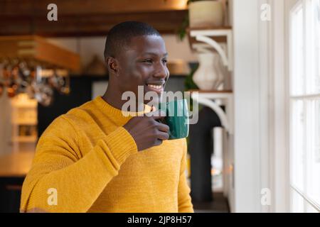 Smiling african american man in kitchen at home drinking coffee and looking out of window Stock Photo