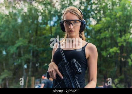 Caucasian woman standing on shooting range looking confidently into camera wearing safety goggles and headphones holding submachine gun. Horizontal shot. High quality photo Stock Photo