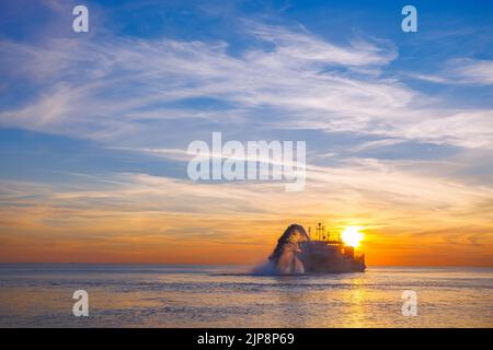Vessel engaged in dredging at sunset time. Hopper dredger working at sea. Ship excavating material from a water environment. Beautiful sunset, Baltic Stock Photo