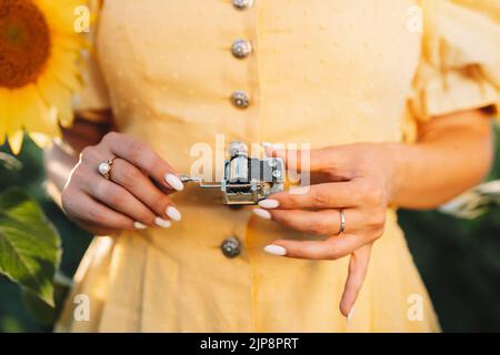 Female hands rotating gears of old music box mechanism. Lady turning the lever of retro small metallic carillon. Woman in sunflowers field listening Stock Photo