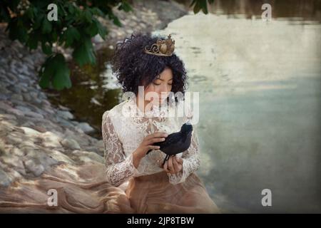 From above of graceful young African American female in princess costume and crown caressing black crow while sitting on rocky shore of calm lake in p Stock Photo