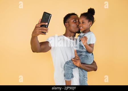 Cheerful african american dad kissing his daughter girl in casual clothes isolated on yellow background studio portrait. People lifestyle concept. Mock up copy space. Doing selfie shot on mobile phone Stock Photo