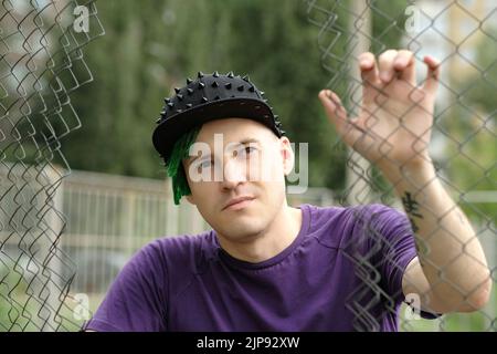 Young man with green dreadlocks in black cap, purple t-shirt posing through hole of lattice fence Stock Photo