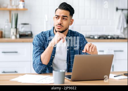 Disappointment upset frustrated indian or arabian young man with glasses, programmer, freelancer, working remotely, sit at home in the kitchen with laptop, looking to the side, sad facial expression Stock Photo