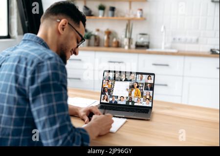 Distance online lecture. Side view of a mixed race guy, male student uses a laptop, on a screen students with a female teacher. The guy studies remotely, listen to lecture, takes notes in a notebook Stock Photo