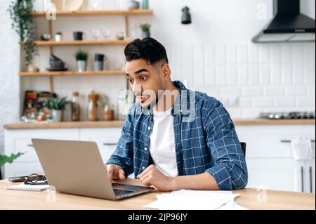 Surprised puzzled mixed race young man, freelancer or creative manager, sitting at home in the kitchen, shocking looking at the laptop screen, in stress of unexpected news, opened his eyes wide Stock Photo
