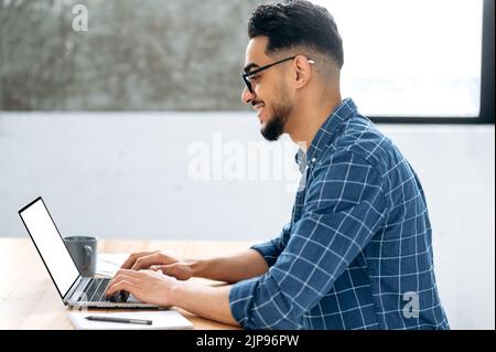 White laptop screen, mockup concept. Arabian or Indian young man, stylishly modernly dressed, programmer, freelancer, sitting at the table with a laptop, typing on the keyboard Stock Photo