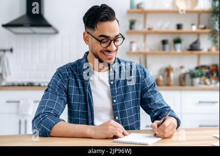 Clever excited Arabian or Indian modern guy, clever student or freelancer, with glasses, listening to an online webinar, lecture, writing down information, gain knowledge, smiling friendly Stock Photo