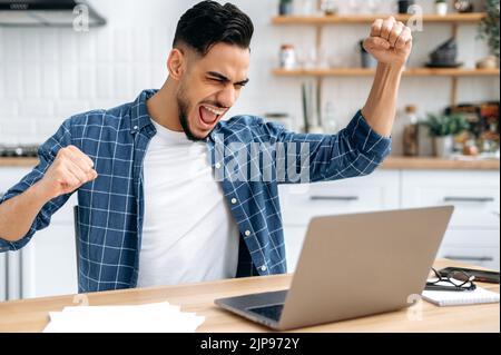 Emotional amazed happy indian or arabian young man, trader, student, freelancer, sitting at table with laptop in the kitchen, enjoying big profit, victory, good news, gesturing with fists, smiling Stock Photo