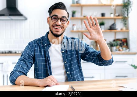 Confident positive indian or arabian man, sit at desk at home, talk on webcam, having video call or conversation with client or colleague, mentor record training course, waves hand, smiling friendly Stock Photo
