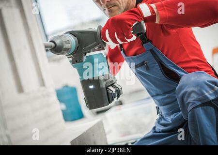 Caucasian Worker Drilling the Hole in the Wall of Newly Built Residential House Using Power Drill. Construction Site in the Background. Industrial The Stock Photo