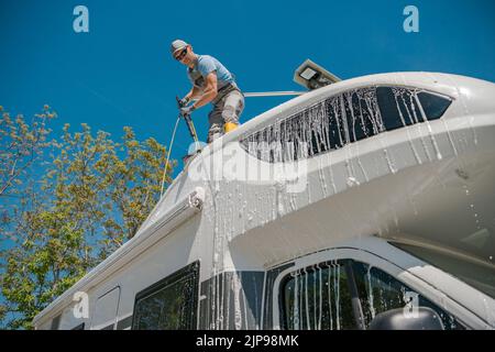 Caucasian Man Washing His White Motorhome While Standing on the Top of Its Roof. Camper Van Owner Taking Care of His Vehicle After a Long Trip. Stock Photo