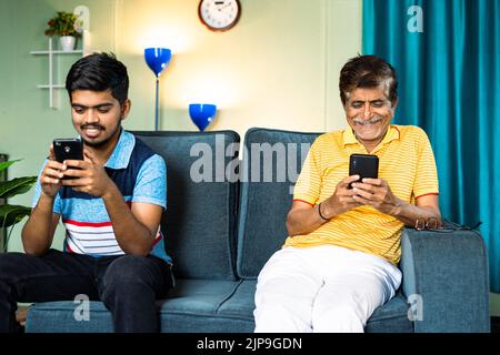 Focus on father, father and son busy using smartphone while sitting on sofa in distance at home - concept of Mobile phone addiction, digital Stock Photo