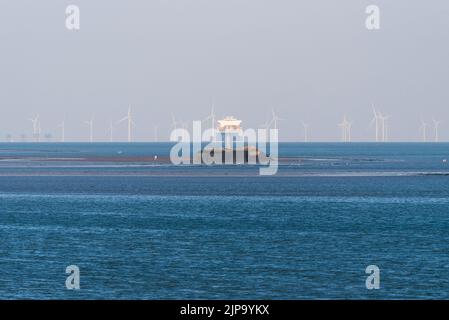 Distant cargo ship navigating through offshore windfarms in Thames Estuary, with walkers far out from shore at Mulberry Harbour section off Southend Stock Photo