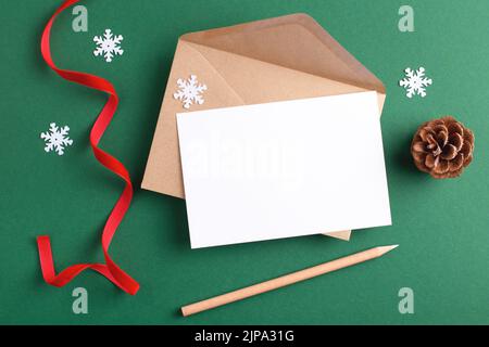 Season's greeting mockup with blank card, kraft envelope, wooden pencil and Christmas decoration on green background. Stock Photo