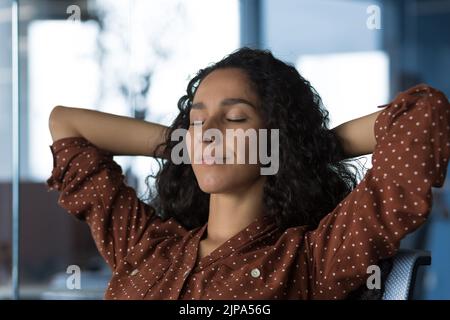 Close-up photo portrait of young beautiful curly Arab woman resting indoors at home, woman with hands behind head resting sitting on sofa near window, eyes closed breathing, meditating and dreaming Stock Photo
