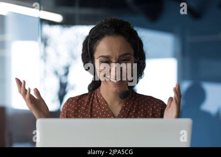 Close-up photo of young beautiful curly Arab woman talking on video call, looking at laptop web camera and smiling using headset for communication, successful businesswoman working in office Stock Photo