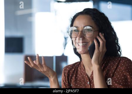 Close-up photo of young beautiful curly Arab woman talking on the phone smiling, business woman in shirt and glasses working in modern office at work Stock Photo